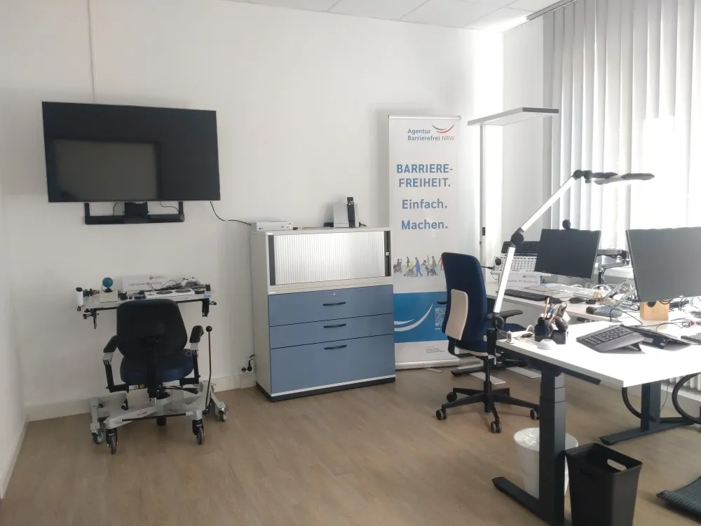 Foto des Demo-Büros in der Ausstellung barrierefreies Arbeiten. Zwei Schreibtische, und ein Fernseher. Darunter ein Therapietisch, daneben ein Büroschrank.