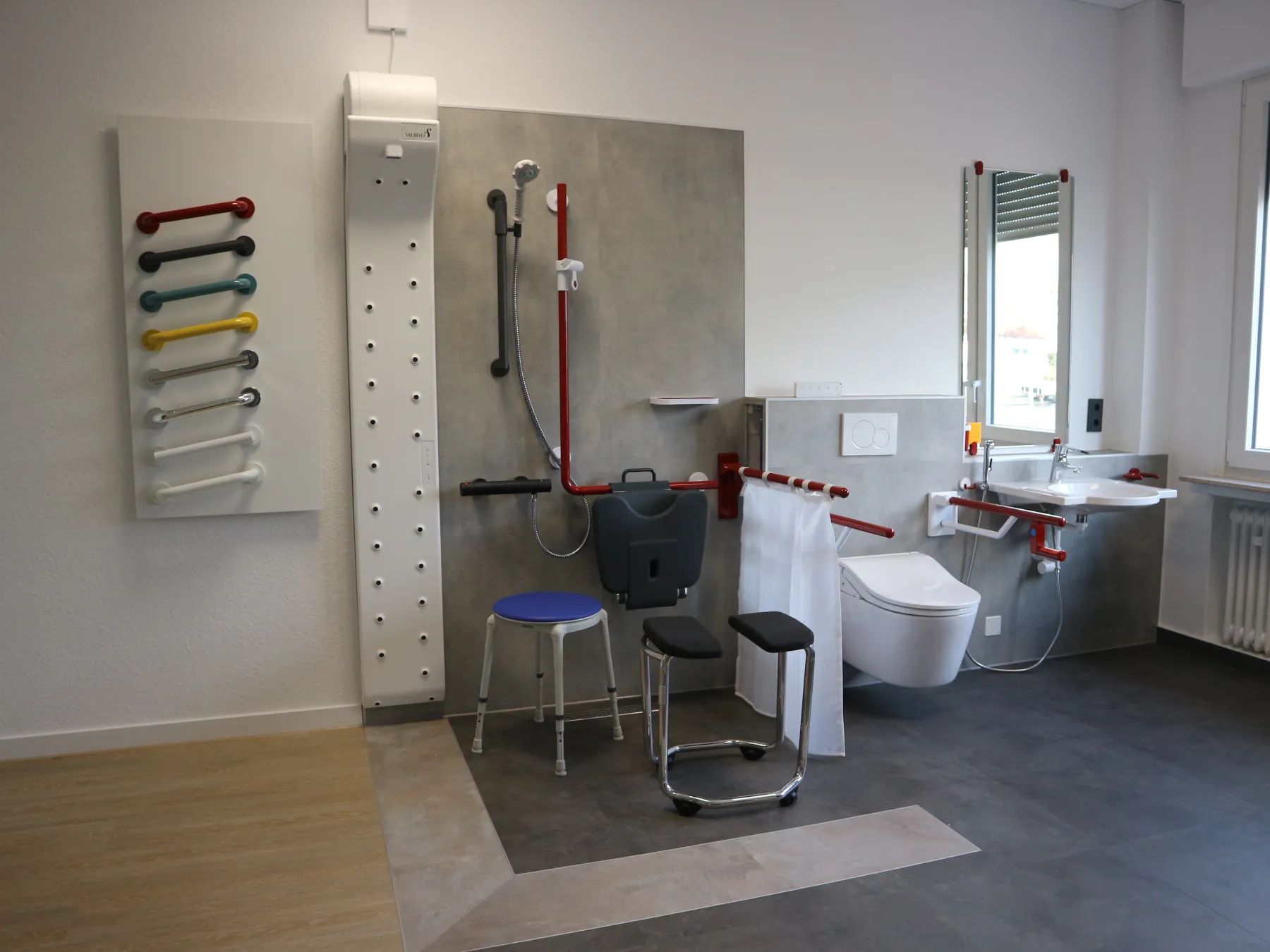 Foto des Ausstellungsbereichs Badezimmer. Eine ebenerdige Dusche, diverse kontrastreiche Haltegriffe, ein Körperföhn, ein Dusch-WC und ein unterfahrbares Waschbecken