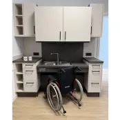 Bild einer höhenverstellbaren und unterfahrbarer hellen Küche mit anthrazid farbener Arbeitsplatte. Der Oberschrank und die Spüle sind leicht nach unten gefahren. Davor ein Rollstuhl.