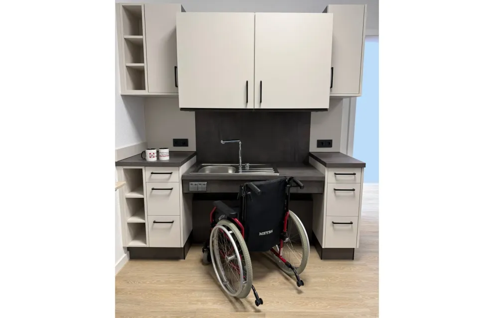 Bild einer höhenverstellbaren und unterfahrbarer hellen Küche mit anthrazid farbener Arbeitsplatte. Der Oberschrank und die Spüle sind leicht nach unten gefahren. Davor ein Rollstuhl.