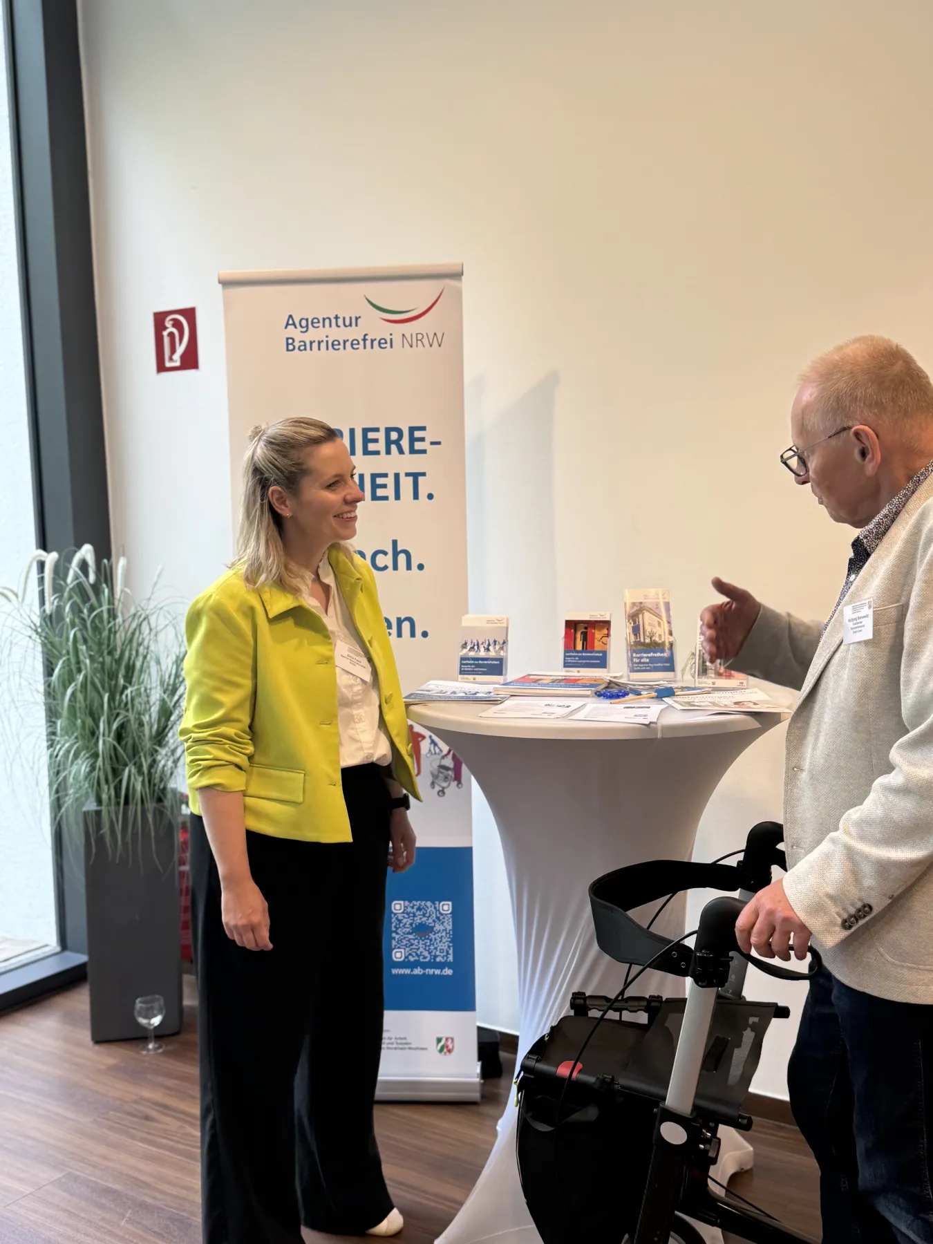 Daria Frank im Gespräch mit einem Mann mit Rollator am Informationstisch der Agentur Barrierefrei NRW auf dem Fachtag für alle