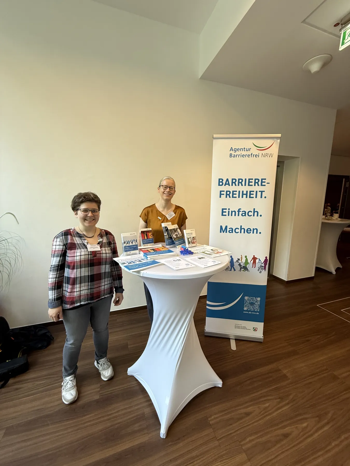 Virginia Grossek und Annika Nietzio vor dem Informationstisch der Agentur Barrierefrei NRW auf dem Fachtag für alle
