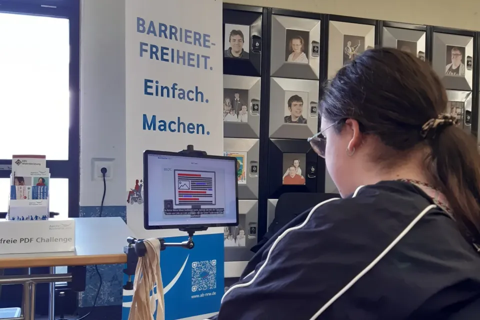 Eine Besucherin erprobt das Spiel Tom’s barrierefreie PDF Challenge. Im Hintergrund das Banner der Agentur Barrierefrei NRW mit dem Text Barrierefreiheit Einfach Machen