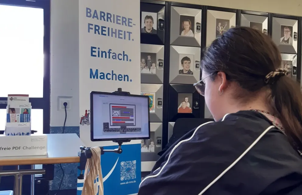 Eine Besucherin erprobt das Spiel Tom’s barrierefreie PDF Challenge. Im Hintergrund das Banner der Agentur Barrierefrei NRW mit dem Text Barrierefreiheit Einfach Machen