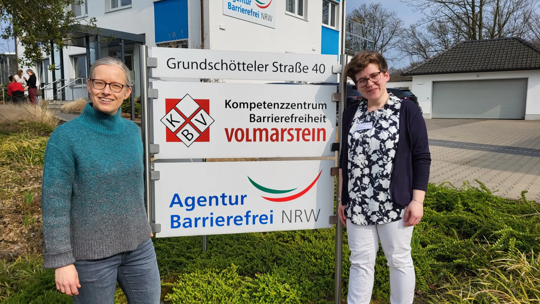 Foto von Annika Nitzio und Virginia Grossek vor dem Hinweisschilds des KBV und der Agentur Barrierefrei NRW