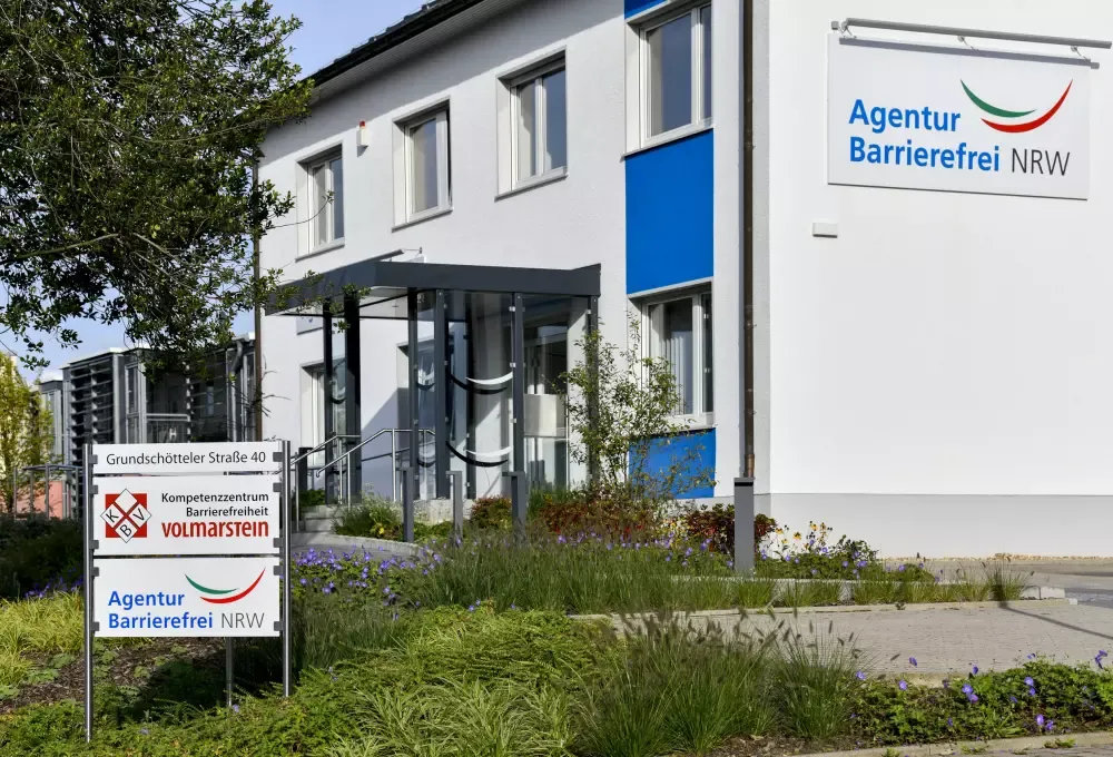 Foto des Gebäudes mit einem Schild KBV und Agentur Barrierefrei NRW an der Einfahrt zum Parkplatz. Am blau weiß gestalteten Gebäude befindet sich ein Logo der Agentur Barrierefrei NRW.