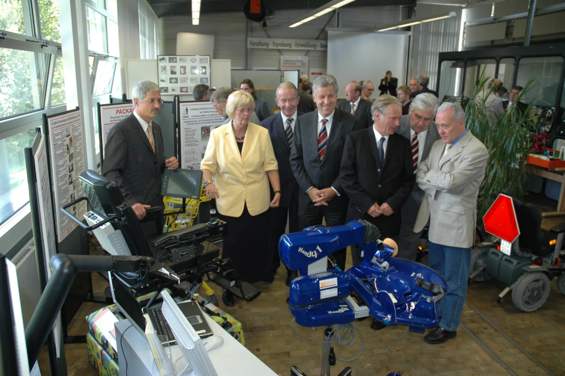 Professor Bühler führt die Besucher begleitet durch Herrn Kaufmann durch die Hilfsmittelausstellung, im Vordergrund das Exponat Handy1.