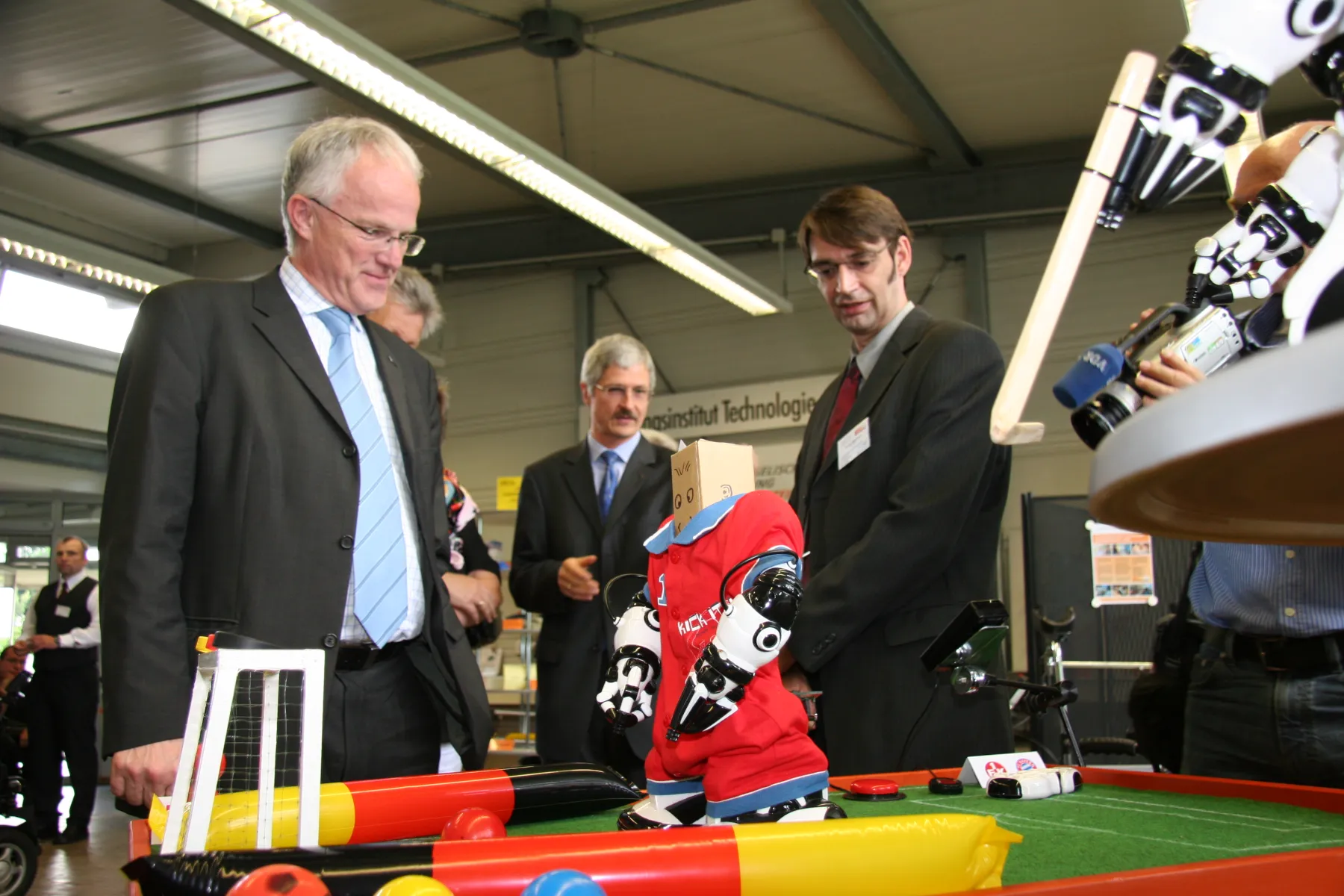 Mitarbeitende des FTB präsentieren Jürgen Rüttgers verschiedene Exponate in der Hilfsmittelausstellung, wie fussballspielende Roboter, im Hintergrund Professor Bühler.