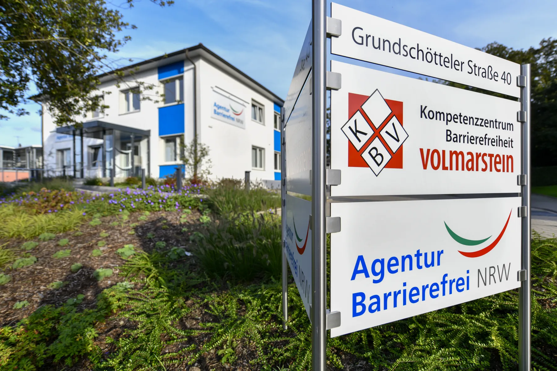 Foto des weiß blau gestalteten KBV-Gebäudes von der Strasse aus gesehen mit Hinweisschildern an der Einfahrt zum Parkplatz, die die Adresse Grundschötteler Strasse 40 enthalten und den Hinweis auf KBV sowie die Agentur Barrierefrei NRW.