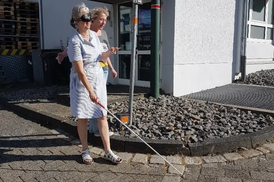 Zwei Frauen laufen eine Rampe runter. Eine Frau hat eine Augenbinde, wodurch die blind ist und hat einen Langstock in der Hand. Die andere Frau sichert sie ab.