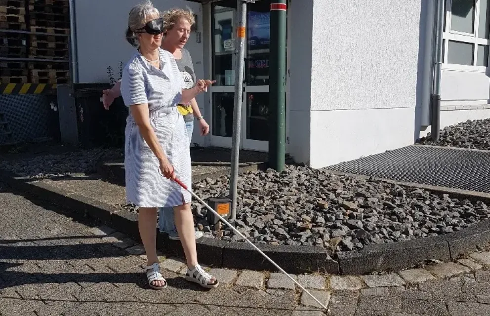 Zwei Frauen laufen eine Rampe runter. Eine Frau hat eine Augenbinde, wodurch die blind ist und hat einen Langstock in der Hand. Die andere Frau sichert sie ab.